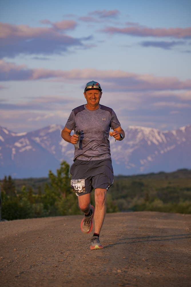 A smiling man running in the dark light of a midnight sun, towards the camera along a road, with snow-capped mountains behind him