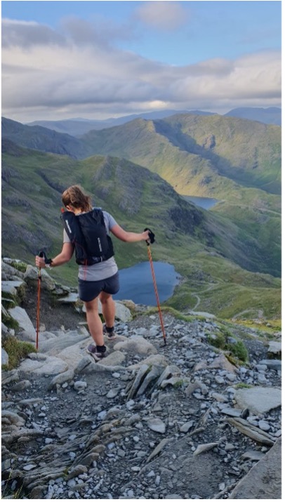 Megan picking her way through rocks and scree at the top of a mountain, with views of lakes and mountains beyond her in her Lakes, Meres and Waters Report