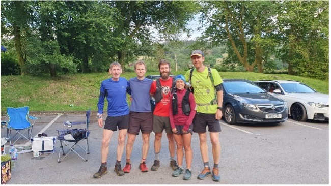 A group of 5 runners standing together with arms round each other in a car park. Megan, significantly smaller than the men around her, looking strong and happy