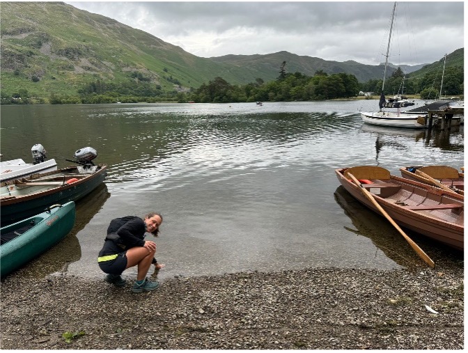 Megan kneeling and touching the waters of a lake, wooden boats either side of her and the lake and a hill behind her in the distance