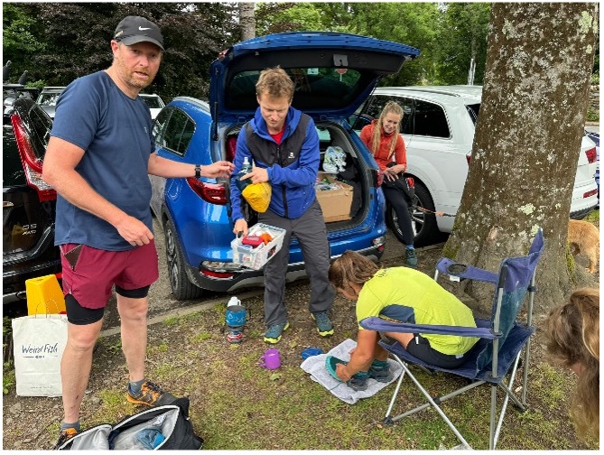 Crew point with cars under trees, Megan sitting in a camping chair and two men offering her food and helping her as she tends to her feet