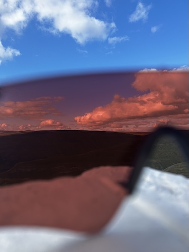 Blurred close-up of a person wearing large sunglasses against a blue sky and orange-pink clouds.