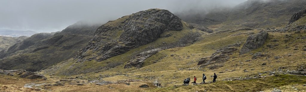 Two runners cross an open, rocky mountain slope beneath dark peaks.