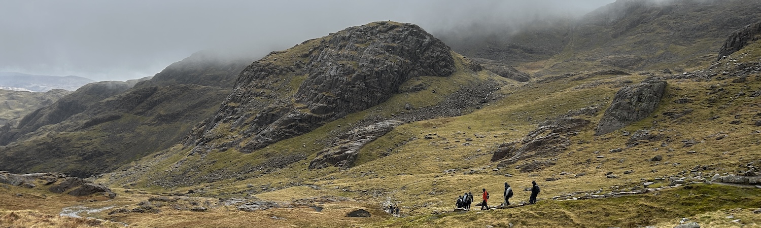 Two runners cross an open, rocky mountain slope beneath dark peaks.