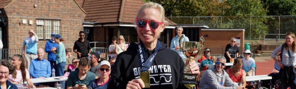 Laura, wearing sunglasses and a race bib, smiles at the camera in front of runners seated around a track.