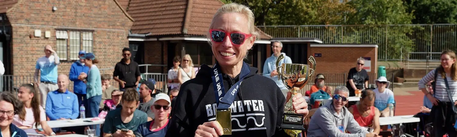 Laura, wearing sunglasses and a race bib, smiles at the camera in front of runners seated around a track.