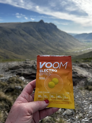 A hand holds a packet of VOOM Electro Bytes against a mountain landscape.